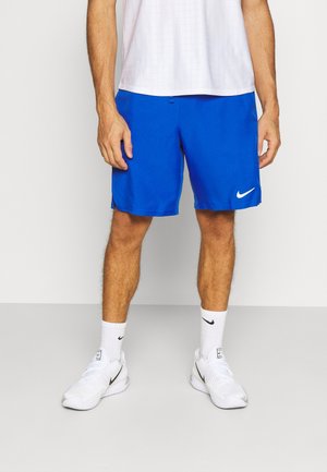 Mann trägt blaue Nike-Shorts, weiße Nike-Socken und weiße Nike-Sneaker und steht vor einem neutralen Hintergrund.
