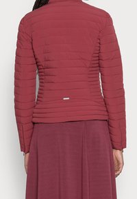 Žena v přiléhavé, prošívané bundě v barvě starorůžové s horizontálním prošitím a odpovídající sukni, zobrazená ze zadní strany.