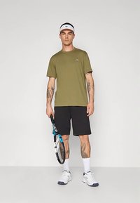 Άνδρας στέκεται κρατώντας ρακέτα τένις, φορώντας φανέλα ελιάς, μαύρο σορτς, λευκά αθλητικά παπούτσια, λευκές κάλτσες και λευκό αθλητικό καπέλο-σκιάδιο.