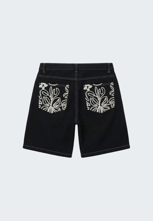 Shorts noirs avec des poches arrière à motif floral blanc et des coutures blanches sur un fond gris clair uni.