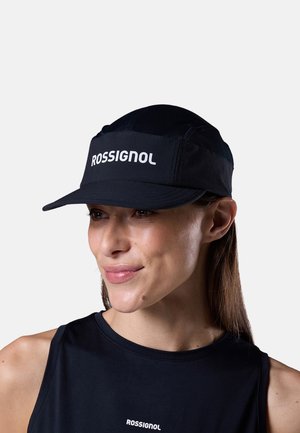 Vrouw die een zwarte Rossignol-pet en een zwarte mouwloze top draagt, glimlacht en kijkt lichtjes opzij tegen een effen achtergrond.