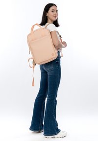 Persikanvärinen reppu, jossa on sileä mattapinta, kaksi yläkahvaa ja säädettävät olkahihnat. Repussa on hillitty logo ja minimalistinen muotoilu.