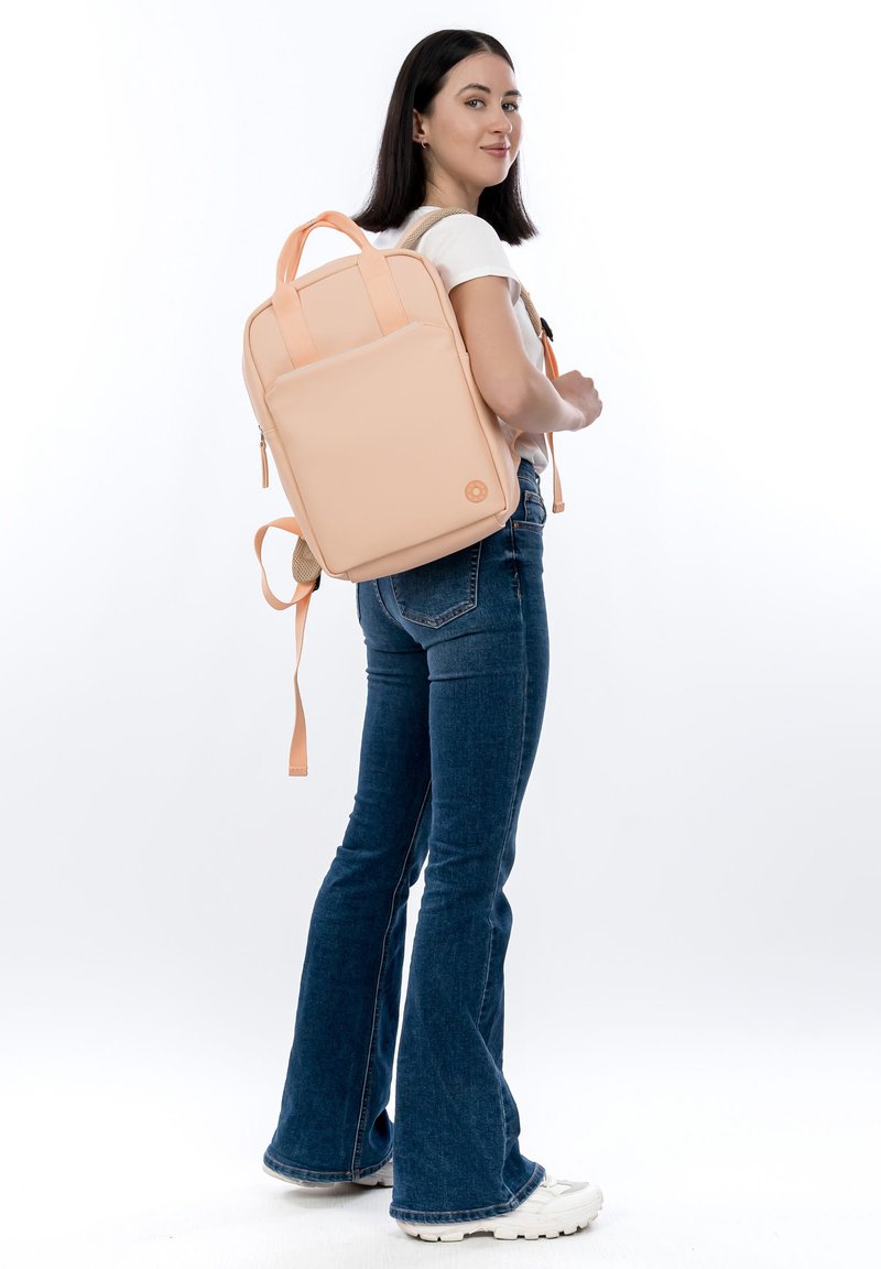 Persikanvärinen reppu, jossa on sileä mattapinta, kaksi yläkahvaa ja säädettävät olkahihnat. Repussa on hillitty logo ja minimalistinen muotoilu.