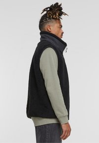 Fekete fleece mellény magas gallérral, cipzáros záródással, és kontrasztos egyszínű ujjak egy világoszöld hosszúujjú póló felett. Laid-back fazon.