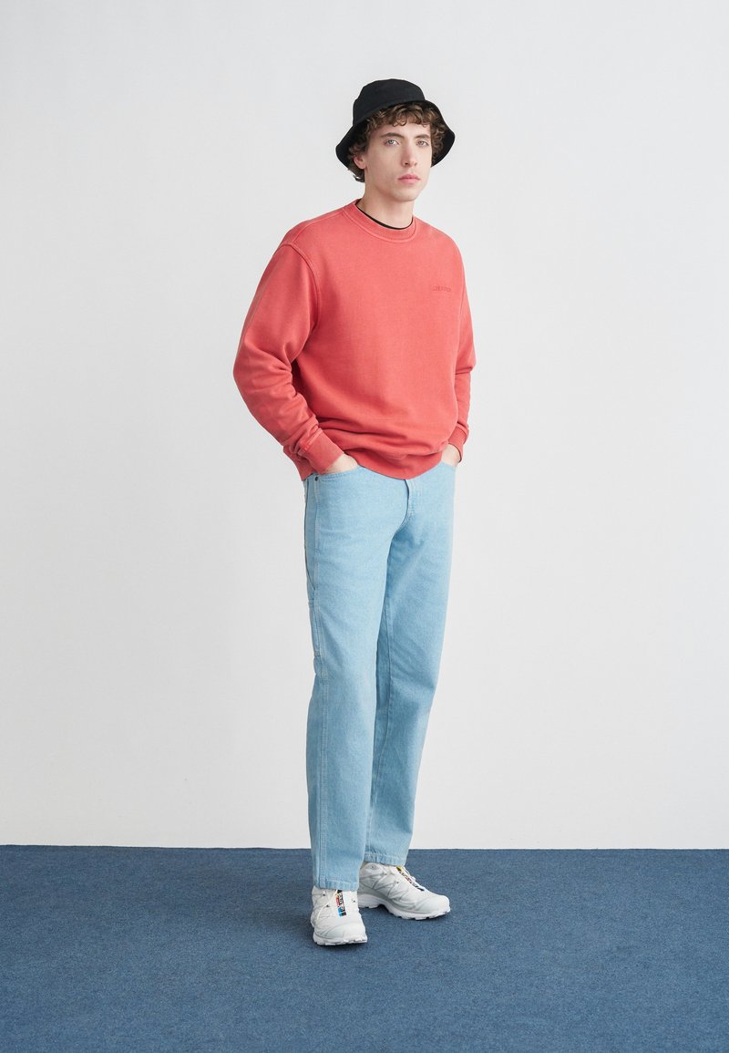 Felpa rossa, jeans azzurri, scarpe da ginnastica bianche e cappello a secchiello nero. Outfit casual con una vestibilità comoda, caratterizzato da una scollatura rotonda.