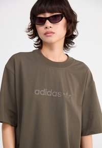 Olive žalia medvilninė marškinė su dideliu pilku adidas logotipu, apvaliu iškirpimu ir laisvo kirpimo rankovėmis. Dėvimi su juodais katės akies akiniais.