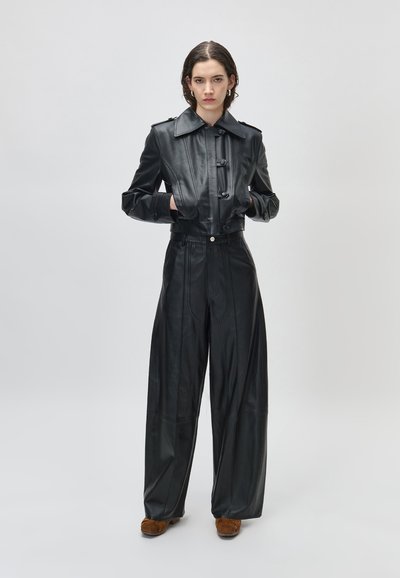 Žena stoji s rukama u džepovima noseći crnu kožnu kratku jaknu, široke kožne hlače i smeđe cipele na jednobojnoj pozadini.