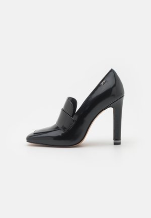 Blank sort loafer med høj hæl, spids tå og remdetalje, med blokhæle og DKNY-logo nær hælen.