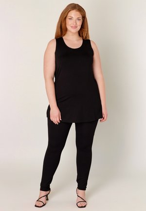 Glimlachende roodharige vrouw die een mouwloze zwarte tuniek, zwarte legging en zwarte sandalen met hoge hakken draagt, staande tegen een eenvoudige lichte achtergrond.