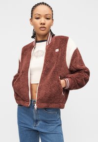 Νεαρό άτομο με πλεγμένα μαλλιά φοράει ένα καφέ και λευκό χνουδωτό μπουφάν, ένα κοντό μπλουζάκι με σχέδιο και μπλε τζιν, στέκεται με τα χέρια στις τσέπες.