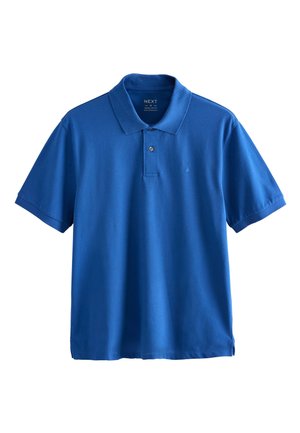 Muž s tetovanými rukami, ktorý nosí modré polo tričko, čierne džínsy a biele tenisky, stojí s jednou rukou v vrecku na jednoduchom pozadí.