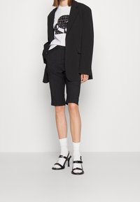 Blazer oversize negro sobre una camiseta gráfica blanca, pantalones cortos negros a medida y tacones negros con tiras y un tacón pequeño. Medias blancas visibles.