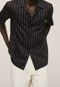 Uomo che indossa una camicia nera a maniche corte con sottili strisce verticali marroni e pantaloni avorio, mani dietro la schiena, su uno sfondo semplice.
