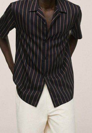 Man wearing black short-sleeve button-up shirt with thin brown vertical stripes and off-white pants, hands behind back, against plain background.