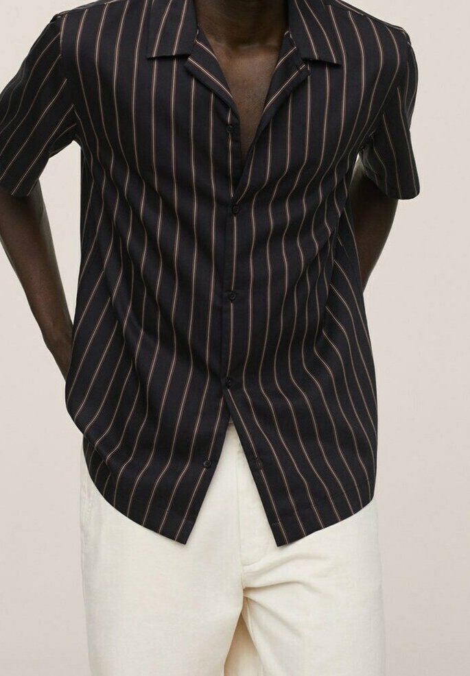 Uomo che indossa una camicia nera a maniche corte con sottili strisce verticali marroni e pantaloni avorio, mani dietro la schiena, su uno sfondo semplice.