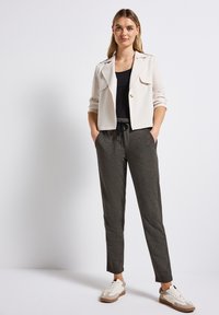 Veste beige coupée sur un top noir, associée à un jogging fuselé gris foncé et des baskets blanches avec des accents marron. Textures lisses tout au long.