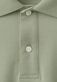Col polo en maille vert clair et patte de boutonnage avec deux boutons blancs, montrant la texture du tissu et les détails des coutures.