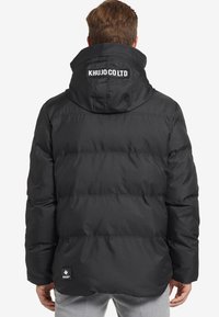 Musta toppatakki hupulla, jossa tikattu pinta, logotunnus takana ja joustavat yksityiskohdat istuvuuden parantamiseksi.