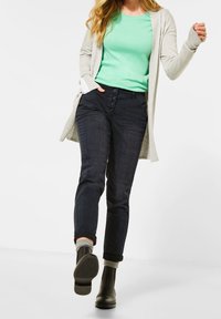 Cardigan gris clair par-dessus un haut vert menthe, assorti à un jean denim foncé avec fermeture à boutons. Bottines noires à la cheville ; ourlets retroussés.
