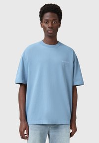 Vaaleansininen puuvillapaita, jossa on rento istuvuus, lyhyet hihat ja pyöreä kaula-aukko. Vasemmassa rinnassa on sävyttä logo.