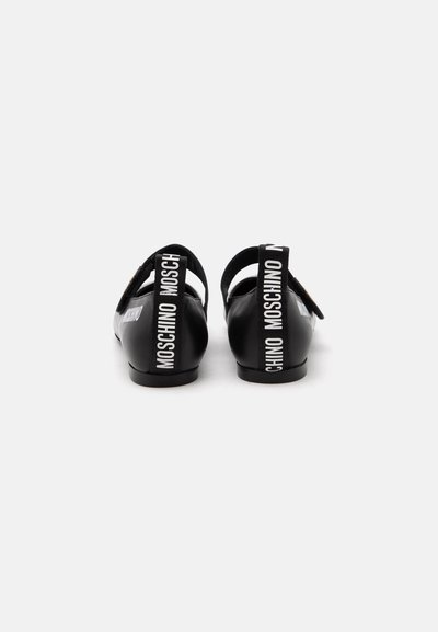 Paire de chaussures plates noires avec brides arrière comportant le texte blanc "MOSCHINO" sur fond blanc.