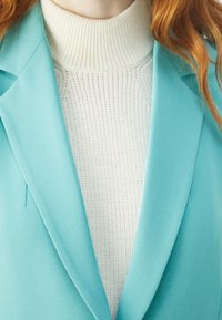 Ušité tyrkysové sako přes bílé žebrované rolák. Sako má ostré klopy a hladkou texturu látky.