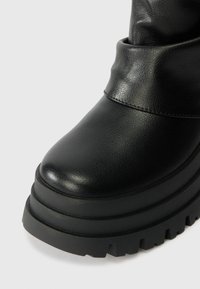 Botín de tobillo de imitación cuero negro con punta redondeada, suela de plataforma gruesa y ribetes cosidos cerca del borde. Parte superior texturizada y suela de goma.