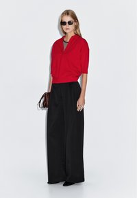 Rode gebreide trui, brede zwarte broek en zwarte zonnebril. De outfit heeft een relaxte pasvorm met een gestreepte binnenlaag en een gestructureerde afwerking.
