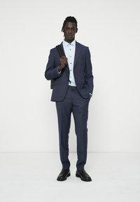 Costum bleumarin, croială ajustată, material texturat, asortat cu o cămașă de culoare bleu deschis și o geantă neagră. Pantofii sunt negri și lustruiți.