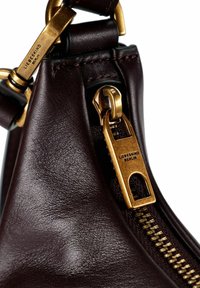 Angolo di borsa in pelle marrone scuro con cerniera e chiusura dorate incise "Liebeskind Berlin" su sfondo bianco.