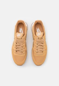 Nike-sneakers med ett beige textilöverdel, runda tår, och sex öglor. Vita logotypaccenster på tungan och sidorna. Lätt texturerad yta.