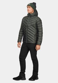 Ciemnozielona kurtka puchowa z pikowanym wzorem, zamek z przodu, połączona z czarnymi spodniami i butami trekkingowymi. Model nosi dzianą czapkę z pomponem.