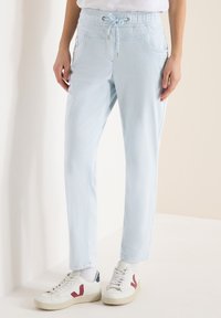 Personne portant un pantalon à cordon de serrage bleu clair et des baskets blanches avec des détails rouges et marines, se tenant devant un fond neutre.