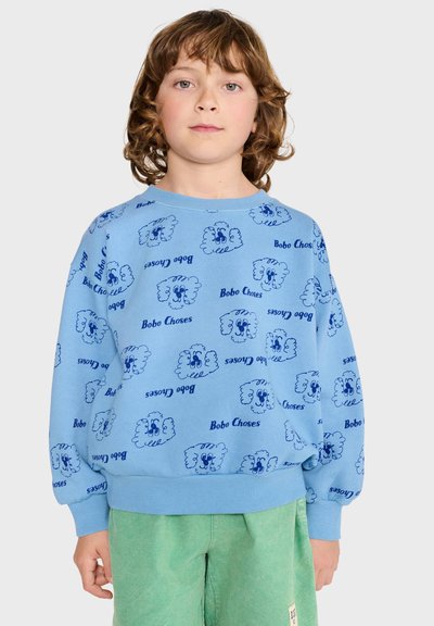 Enfant aux cheveux bouclés portant un sweat-shirt bleu avec des visages de dessins animés et le texte "Bobo Choses", associé à un pantalon vert sur un fond uni.