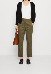 Olijfgroene broek met een elastische tailleband, gecombineerd met een witte t-shirt en een zwarte cardigan, met een bruine leren handtas. Zwarte schoenen.
