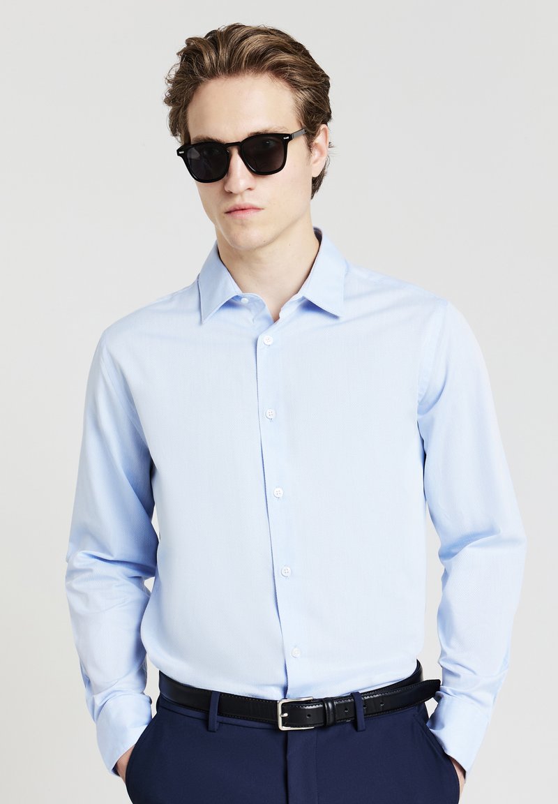 Camicia azzurro chiaro con colletto ben rifinito e una texture sottile, abbinata a occhiali da sole neri e una cintura scura.