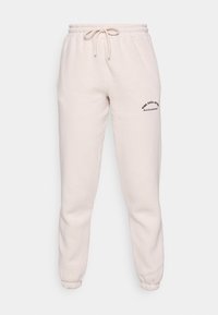 Ljust beige joggingbyxor tillverkade av mjukt material, med elastisk midja och dragsko samt manschetter vid anklarna; vänster sida har tryckt text med logotyp.