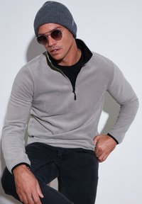 Grå fleecepullover med en kvart-zip krave, båret over en sort skjorte. Modellen har parret den med mørke jeans og en grå strikket hue.