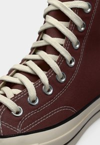 Κοντινό πλάνο μπορντό ψηλού τύπου αθλητικού παπουτσιού με κρέμ κορδόνια, μεταλλικά κουμπώματα, λευκή ραφή και μπεζ λαστιχένια σόλα σε απλό φόντο.