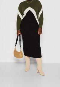 Abito in maglia con maniche lunghe, color blocking verde oliva e nero, caratterizzato da un motivo a zigzag bianco; abbinato a una borsa in tessuto e stivali beige.