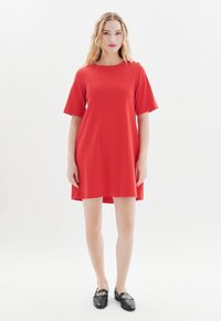 Rotes Kleid mit kurzen Ärmeln, einem runden Ausschnitt und lockerer Passform. Kombiniert mit schwarzen Slippern, die einen goldenen Kettenakzent haben.