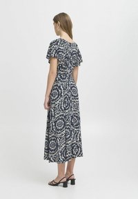 Dunkelblaues Blumenkleid mit weißem abstraktem Muster, kurzen schwingenden Ärmeln, in der Mitte der Wade und kombiniert mit schwarzen Blockabsatzsandalen.