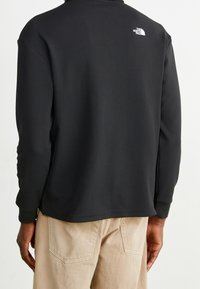 Svart långärmad skjorta med strukturerad tyg, med hög krage och en liten vit logotyp på övre ryggen. Designad för komfort och mångsidighet.