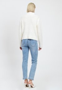 Persoon met kort blond haar, gekleed in een witte gebreide trui, lichtblauwe spijkerbroek en witte hoge hakken, staand met de rug naar de kijker.