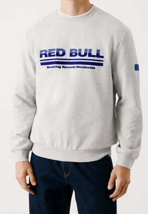 Mann trägt hellgrauen Sweatshirt mit dem Schriftzug „RED BULL Breaking Records Worldwide“ in Blau und dunkelblaue Jeans.