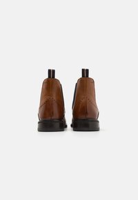 Botas de cuero marrón hasta el tobillo con paneles elásticos laterales en azul marino, presentan un ligero detalle de brogue, suela de goma negra y una lengüeta trasera.