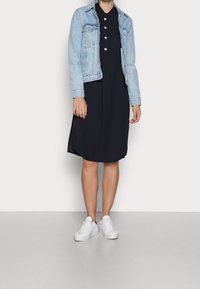 Giacca di jeans azzurra sopra un vestito nero con orlo arrotondato e bottoni. Sneakers bianche completano l'outfit. Texture lisce visibili.