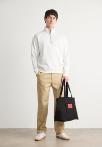Hvitt fleece quarter-zip genser, beige chinos, svart totebag med rød etikett, og hvite sneakers. Enkelt design, avslappet stil, teksturert stoff.