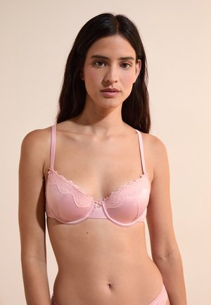 Jonge vrouw die een lichtroze beha met kanten rand draagt, staat tegen een effen beige achtergrond, kijkt recht vooruit met een neutrale uitdrukking.