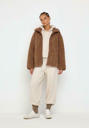 Ung kvinde iført en brun fleecejakke med lynlås, beige hættetrøje, beige træningsbukser, brune sokker og hvide sneakers, der står mod en hvid baggrund.